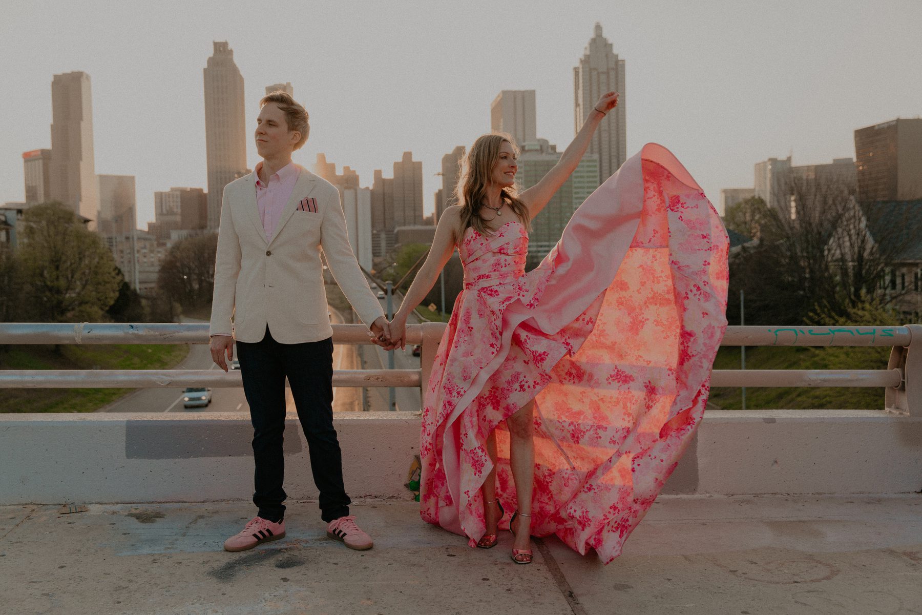 Kimberly and Ian holding hands on a bridge with the Atlanta skyline behind them, her floral gown flowing in the wind