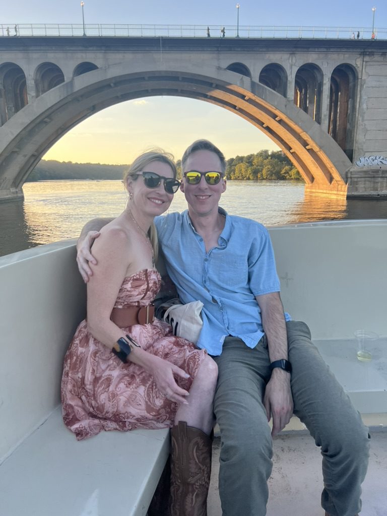 Kimberly and Ian on a boat at sunset in front of a large stone arch bridge
