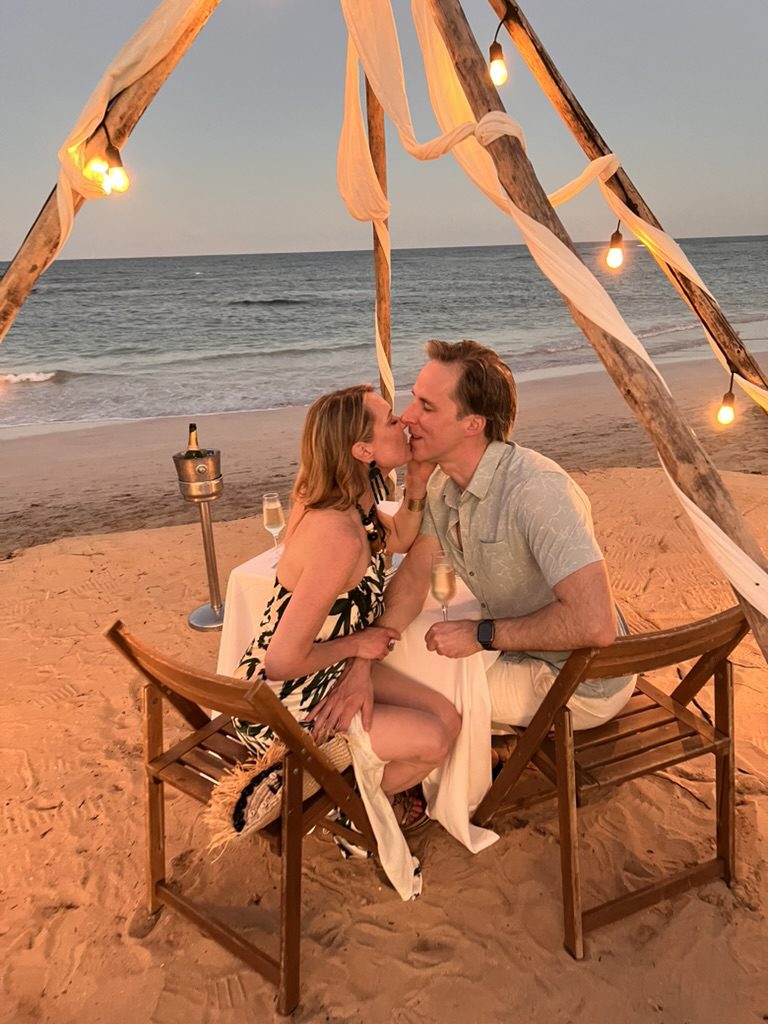 Kimberly and Ian kissing at a candlelit beach table under a canopy of string lights