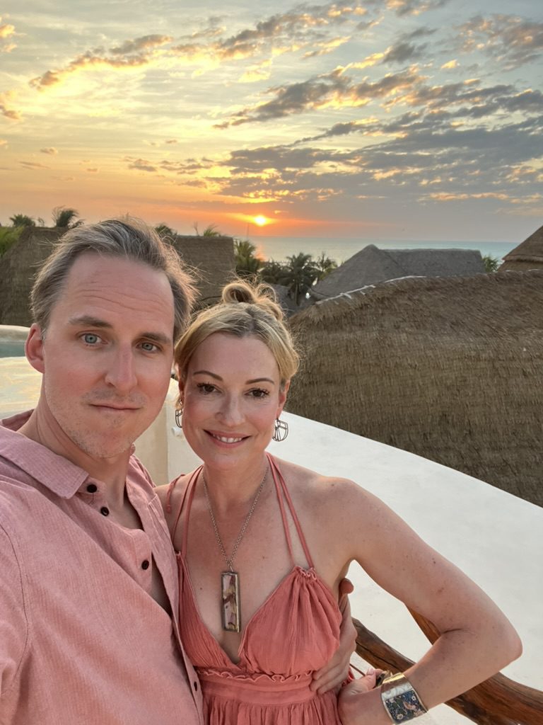 Kimberly and Ian on a rooftop at sunset, with the sun setting over thatched roofs and the ocean
