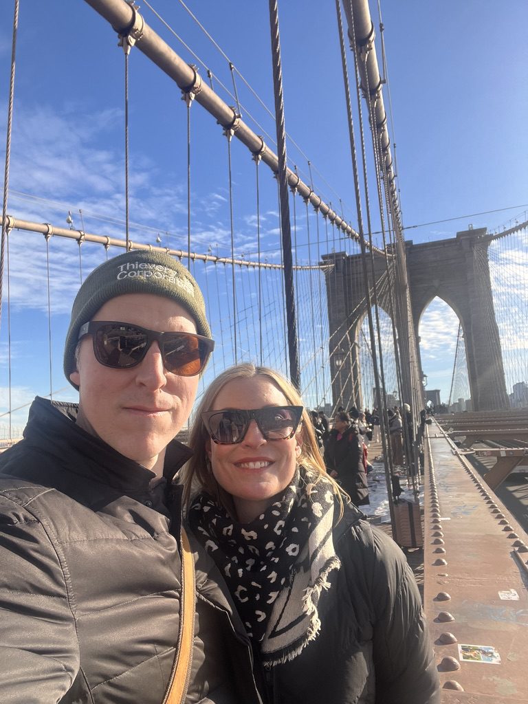 Kimberly and Ian in winter hats and jackets on the Brooklyn Bridge on a sunny day