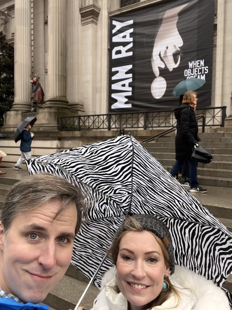 Kimberly and Ian under a zebra-striped umbrella on the museum steps in front of a Man Ray banner