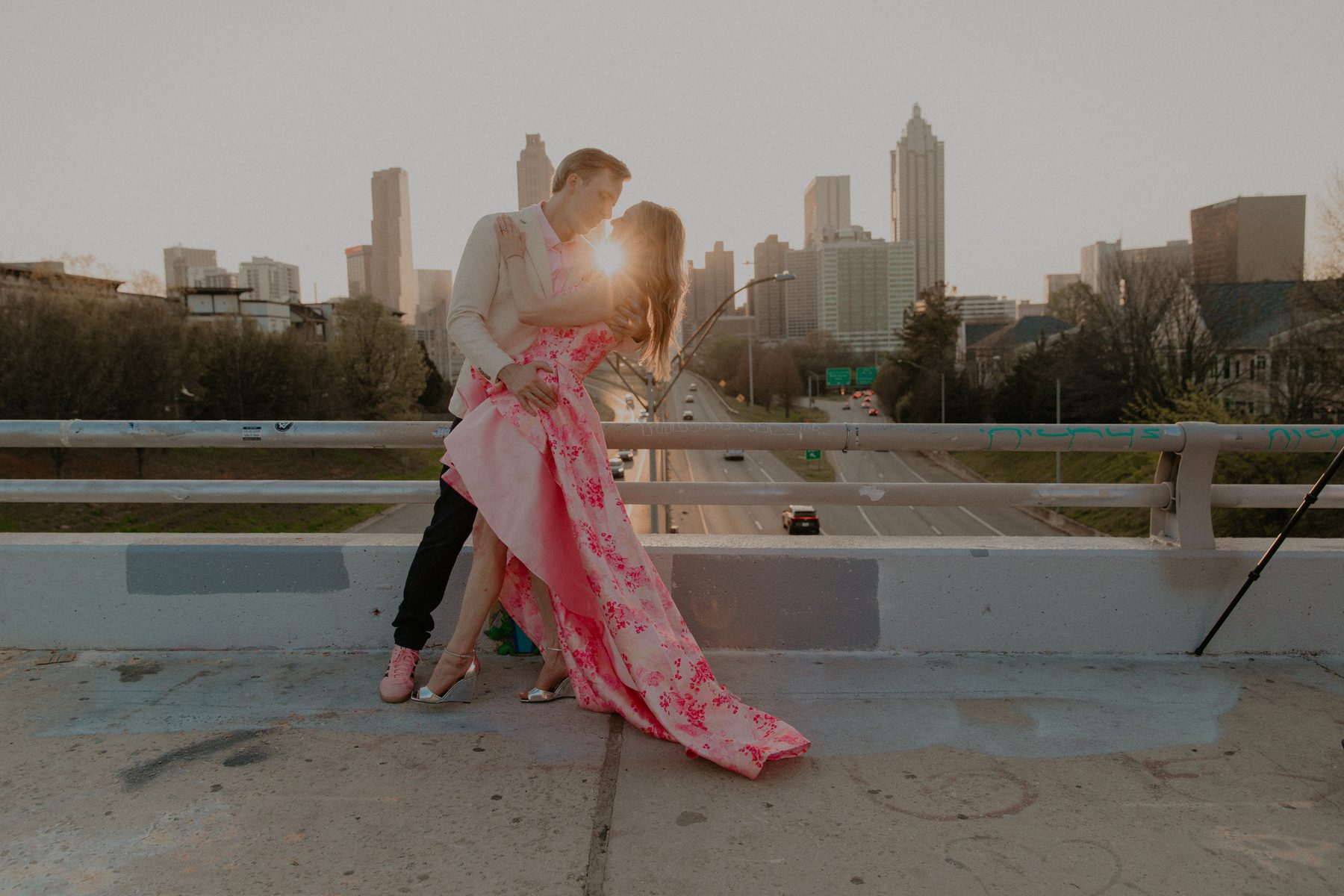 Ian dipping Kimberly for a kiss on a bridge at sunset, the sun flaring between them and the Atlanta skyline behind