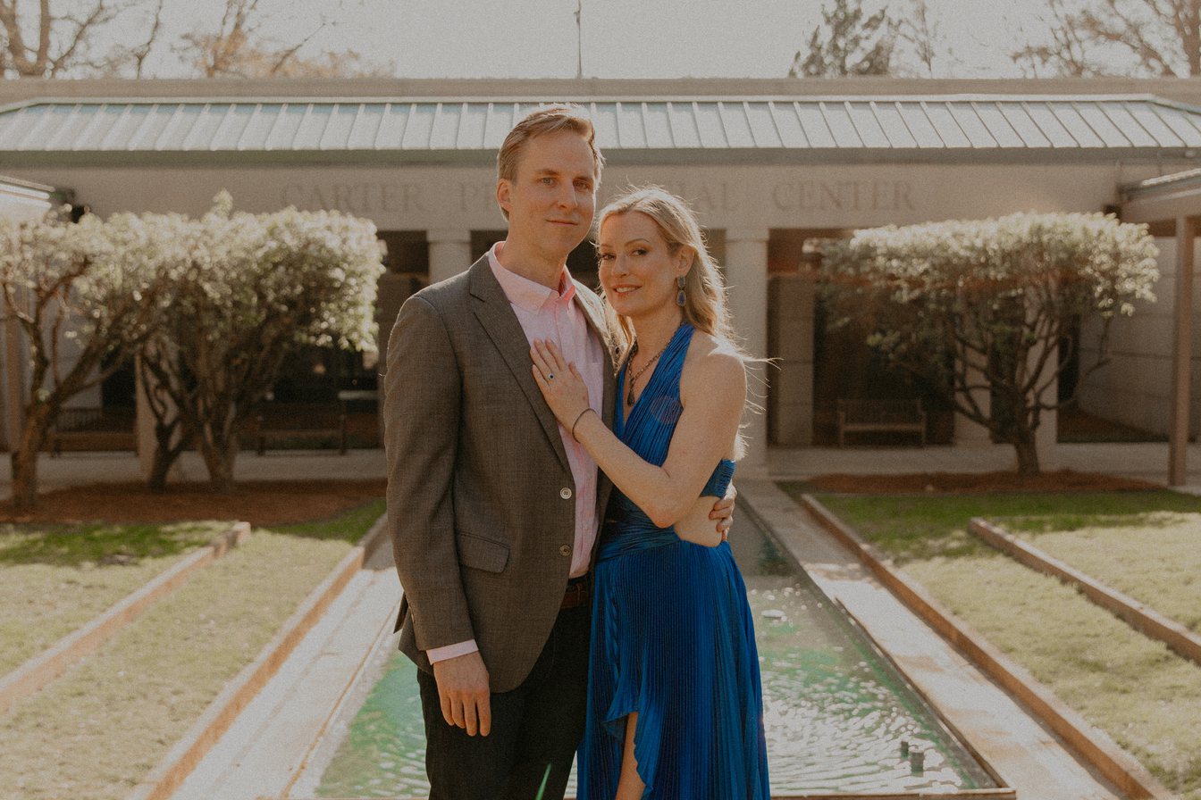 Close-up of Ian in a gray blazer and Kimberly in a blue gown, arm in arm, smiling in front of blooming trees and a reflecting pool